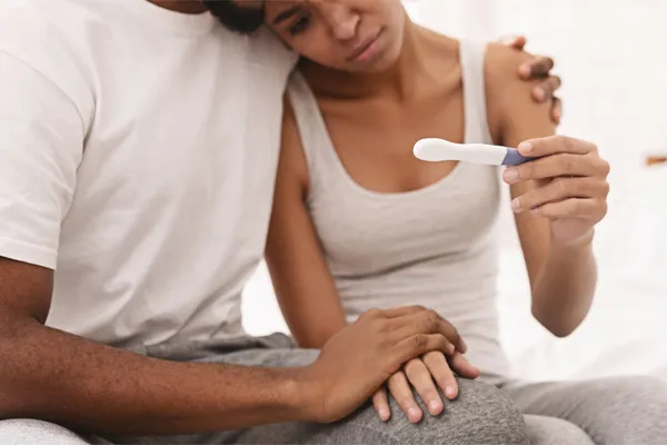 Infertility-Clinic A black couple sits on a bed, disappointed with the negative results of a pregnancy test. Get treatment for infertility from Dr. Lashea Davis Roland in Silver Spring.