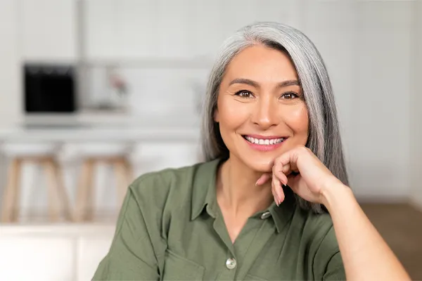 An asian woman with long gray hair and a green shirt sitting and smiling on her couch, happy with her menopause care from [NAME} in Silver Spring.