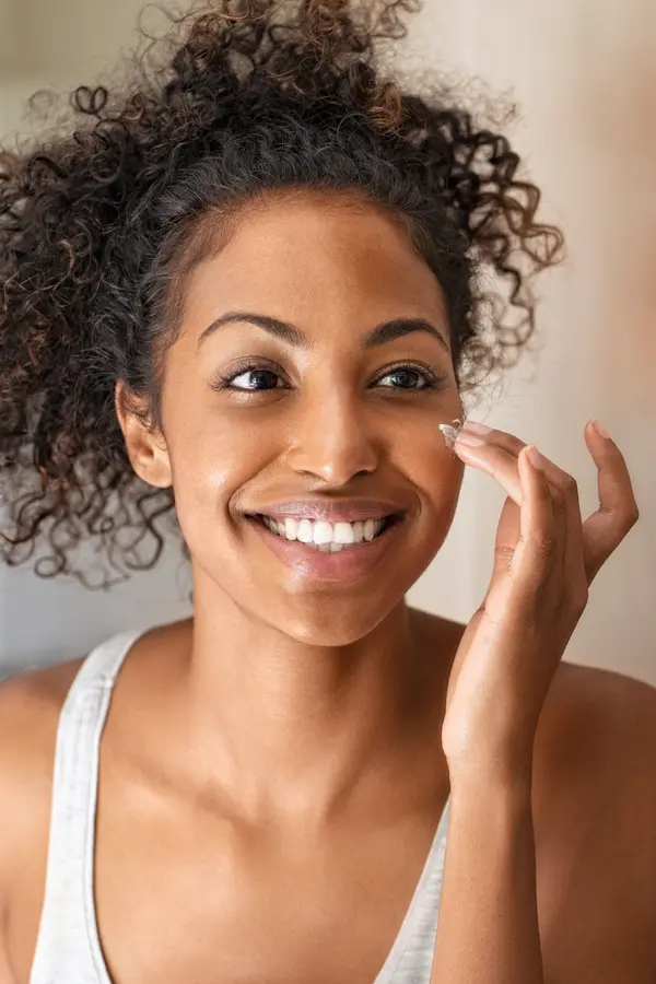 A black woman smiling into the mirror while applying face cream. Excited to have received Morpheus8 Skin Rejuvenation treatment by Dr. Lashea Davis Roland in Silver Spring.