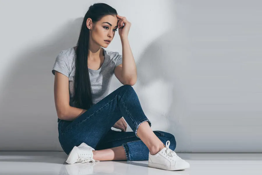 A young brunette woman in a gray t-shirt and jeans sits against a gray wall with her head resting on her hand and a distressed expression. Get help for Polycystic Ovarian Syndrome (PCOS) from Dr. Lashea Davis Roland in Silver Spring.